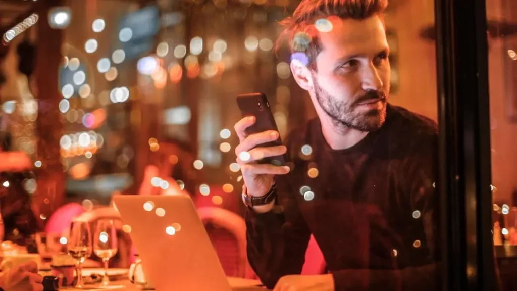 Stylish man using smartphone and laptop at Budapest cafe enhances guest experience with mobile charging solutions.