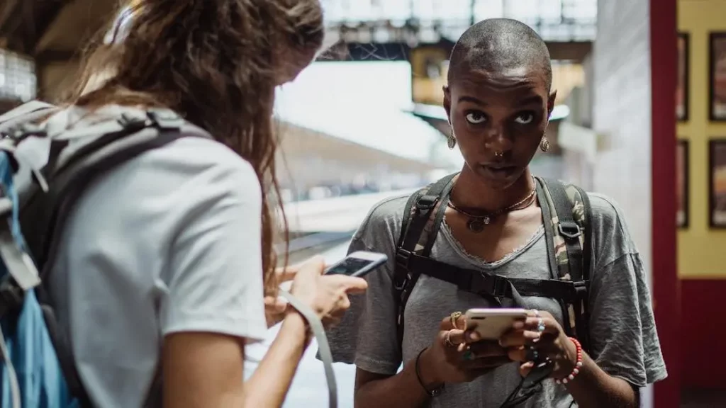 Female travelers using smartphones near Phone Charging Stations at a train station with visible backpacks.