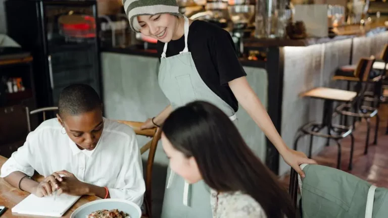 Asian waitress serves pasta to diverse customers in cafe, highlighting "Convenience Drives Repeat Visits".